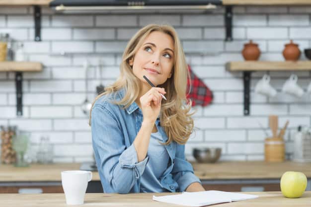 Woman in a kitchen daydreaming, holding a pen with a notebook, coffee cup, and apple on the table in front of her.