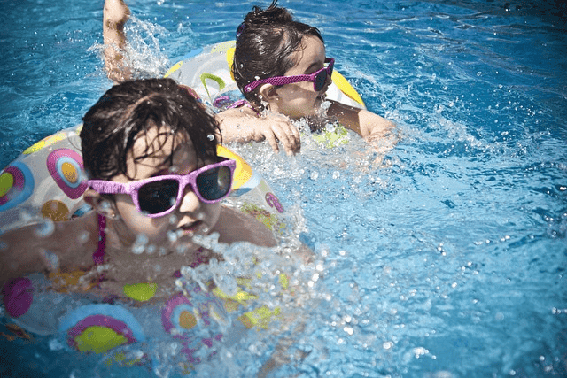 Kids enjoying a sunny day in the pool with colorful floaties and pink sunglasses, splashing water excitedly.