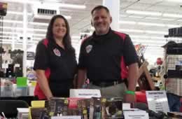 Two people standing behind a display table with various products at a trade show in a brightly lit venue.