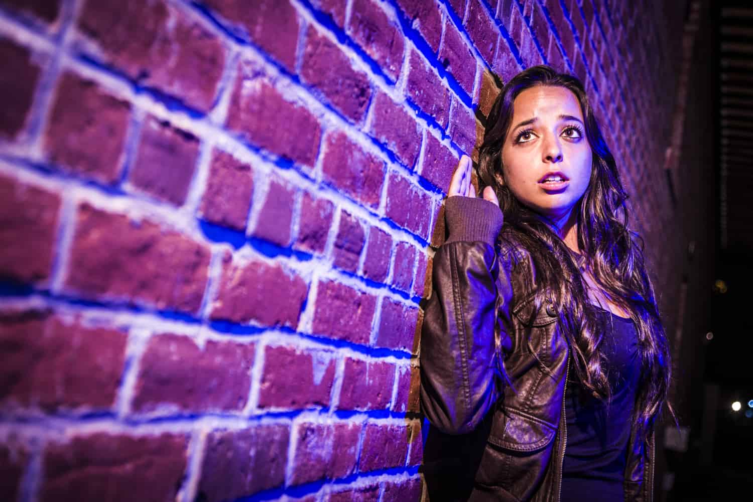 Woman in leather jacket leans against a brick wall under dramatic blue and pink lighting.