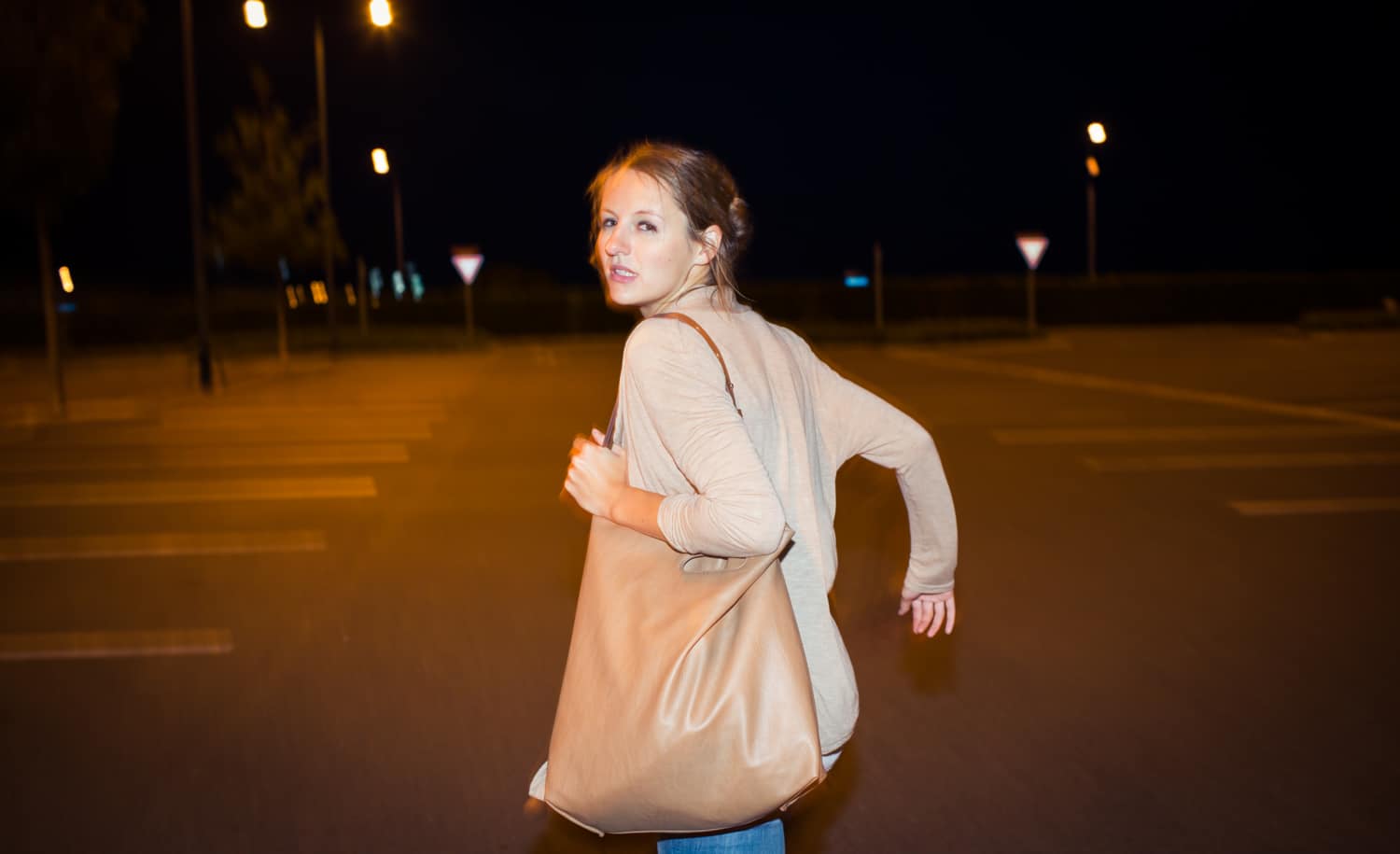 Woman with a tan handbag walking at night in an illuminated parking lot, glancing back over her shoulder.