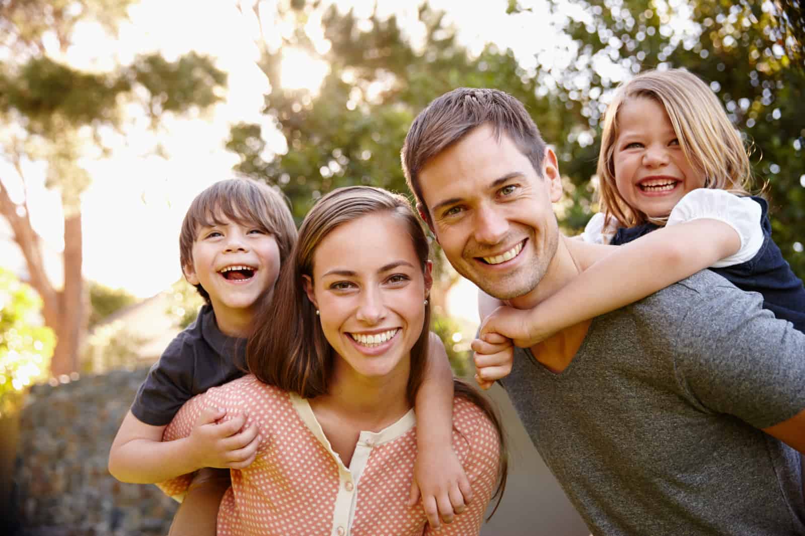 Happy family enjoying time together outdoors, smiling parents with children on their backs in a sunny garden setting.