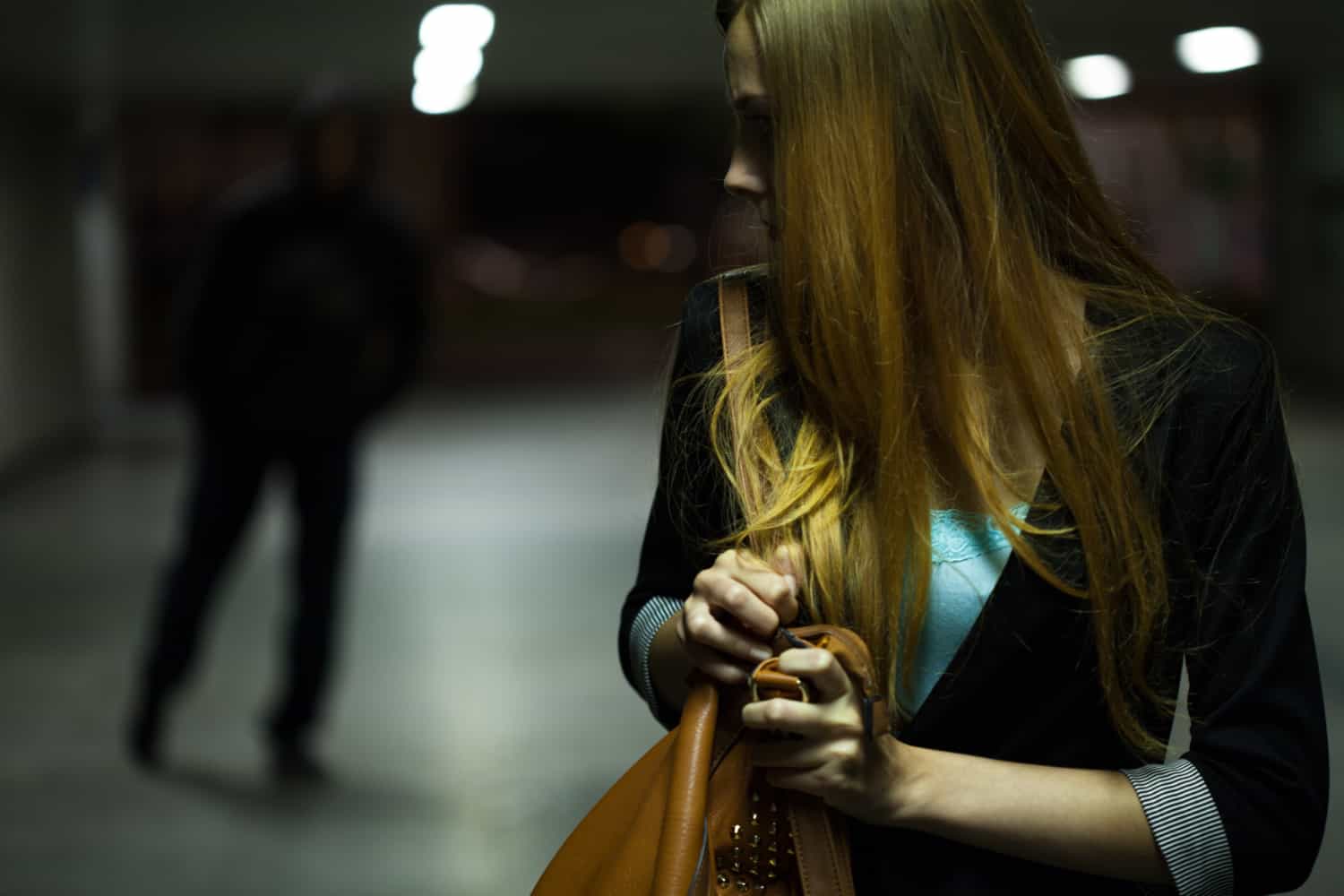 Woman looking back in a dimly lit hallway with a person in the background, holding her handbag tightly.